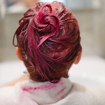 an elementary school age girl dyes her hair pink with a white towel over her shoulders in a gray bathroom near the sink and mirror back view, focus on hair