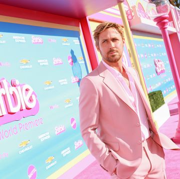 ryan gosling, wearing a pink suit jacket, shirt, and pants, standing in front of a display for the film barbie